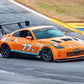 An orange sports car with the number 77X and T2 decals speeds around a racetrack, flaunting its 9livesracing 350z Big Wang Kit '02-08 Z33 performance. The car features extruded aluminum wings, black wheels, and various sponsor stickers. The track is gray with a yellow and blue striped barrier on the side, and grass is visible in the background.