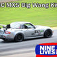 A silver Mazda MX-5 race car with the number 857 and various sponsor stickers on its body is driving on a racetrack. The car, featuring a Miata Big Wang Kit '06-15 NC *V1* aluminum wing from 9livesracing, showcases the text "NC MX5 Big Wang Kits" at the top and a "9livesracing" logo at the bottom.