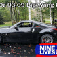 A black Nissan 350z with a large rear wing parked on a wet road surrounded by trees. The text above the car reads, "350z Big Wang Kit '02-08 Z33." Featuring extruded aluminum wings for superior GT3 performance. A blue and white logo at the bottom right corner says "9livesracing.
