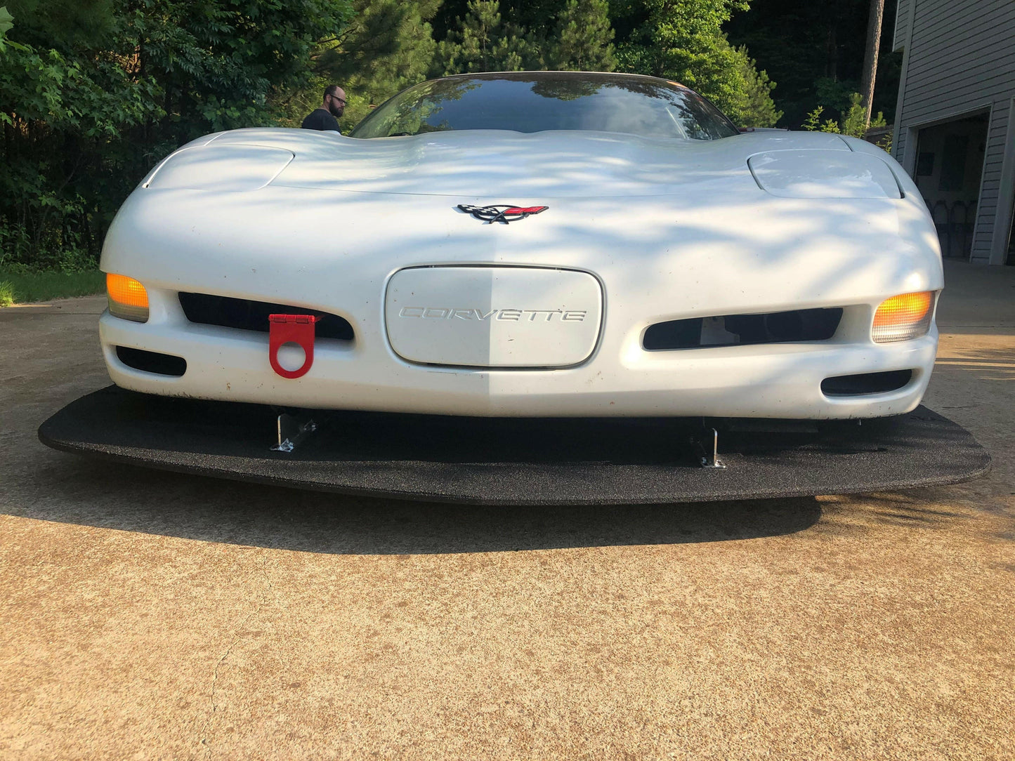 A white Corvette fitted with a 9livesracing Corvette Rocket Nose Splitter ’97-04 C5 is parked on a concrete driveway. The car features a red tow hook attached to the grille area. Trees and a building are visible in the background, completing the sleek look of this performance-oriented machine.