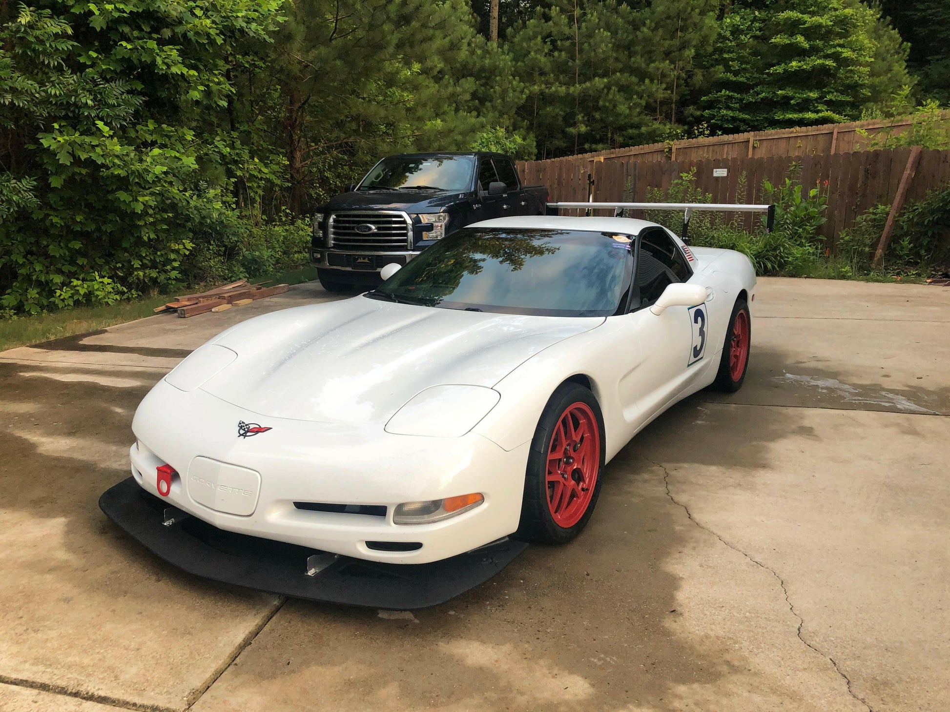 A white sports car with red rims, a racing number "3" on the side, and a black Corvette Rocket Nose Splitter ’97-04 C5 by 9livesracing for added downforce is parked on a concrete driveway. Behind it is a black pickup truck. Trees and a wooden fence are visible in the background.