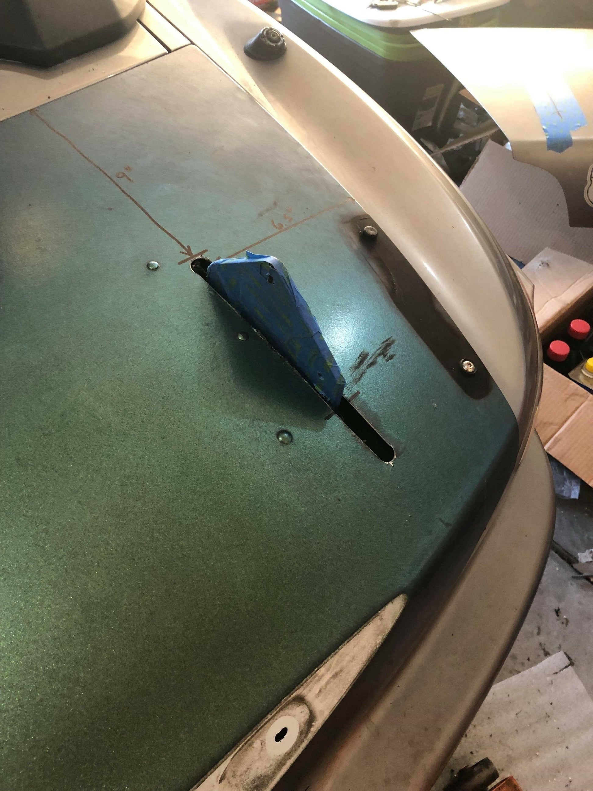 Close-up of a dashboard in progress for a Honda S2000, showing green material being cut with a blue utility knife. Pencil marks and measurements are visible on the surface. The surrounding area appears cluttered with various tools and items, reflecting the meticulous attention to detail needed for optimal down-force adjustments when installing the 9livesracing S2000 Big Wang Chassis Mount Kit '99-09 AP1/AP2.