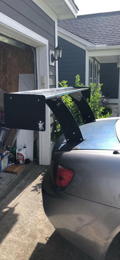 A 9livesracing S2000 Big Wang Chassis Mount Kit '99-09 AP1/AP2, a large, black spoiler with an exaggerated design optimized for down-force, is attached to the rear of a silver Honda S2000 parked in a driveway. The open garage next to the car contains various tools and equipment. A sticker depicting a small, simplified human figure is on the spoiler.