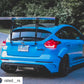 A bright blue Ford Focus RS, equipped with the 9livesracing Focus/RS Big Wang Kit '11-18 3rd Gen aluminum wings for added down-force, is parked on the side of the road. The car features an oversized rear wing, a widebody kit, and sporty decals, including the number "003" on the side. It's situated against a backdrop of trees and greenery.