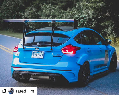 A bright blue Ford Focus RS, equipped with the 9livesracing Focus/RS Big Wang Kit '11-18 3rd Gen aluminum wings for added down-force, is parked on the side of the road. The car features an oversized rear wing, a widebody kit, and sporty decals, including the number "003" on the side. It's situated against a backdrop of trees and greenery.