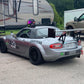 A gray race car, labeled with number 857, is parked in front of an RV and a van in a paddock area. The car features a Miata Big Wang Kit '06-15 NC *V1* by 9livesracing, black wheels, and racing decals, including a Mazda logo. A helmet rests on the car's roof.