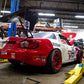 A red and white racing car equipped with the 9livesracing Miata Big Wang Kit '90-'05 NA/NB is elevated on a hydraulic lift in an American-made workshop garage. The car boasts a large rear wing for increased downforce and red rims. Various tools and parts are visible in the background, with shelves and other equipment scattered throughout the space.