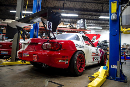 A red and white racing car equipped with the 9livesracing Miata Big Wang Kit '90-'05 NA/NB is elevated on a hydraulic lift in an American-made workshop garage. The car boasts a large rear wing for increased downforce and red rims. Various tools and parts are visible in the background, with shelves and other equipment scattered throughout the space.