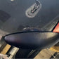 Close-up view of the Miata Headlight Blanks NB1-Only by Nine Lives Racing on a black vehicle, showing part of the mounting area crafted from robust plastic and an attached wiring component. The surrounding area includes reflections and portions of the car's body, highlighting the vehicle's glossy finish.