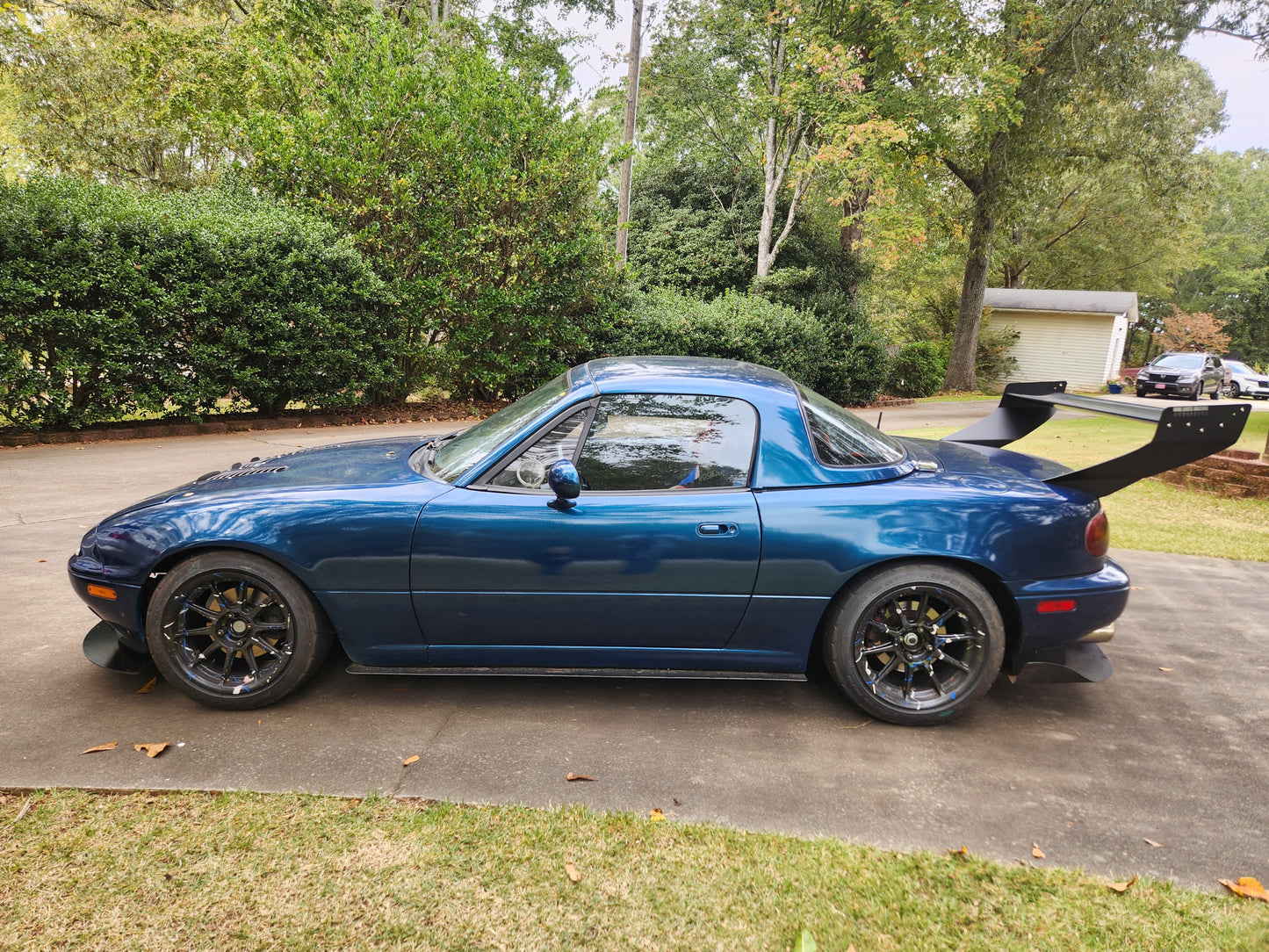 A blue convertible sports car featuring the Miata V2 Street Wang '90-05 NA/NB by 9livesracing is parked on a driveway. The car showcases black alloy wheels and is surrounded by greenery, with a white building in the background. Its performance-oriented design is accentuated by the presence of the impressive rear spoiler.