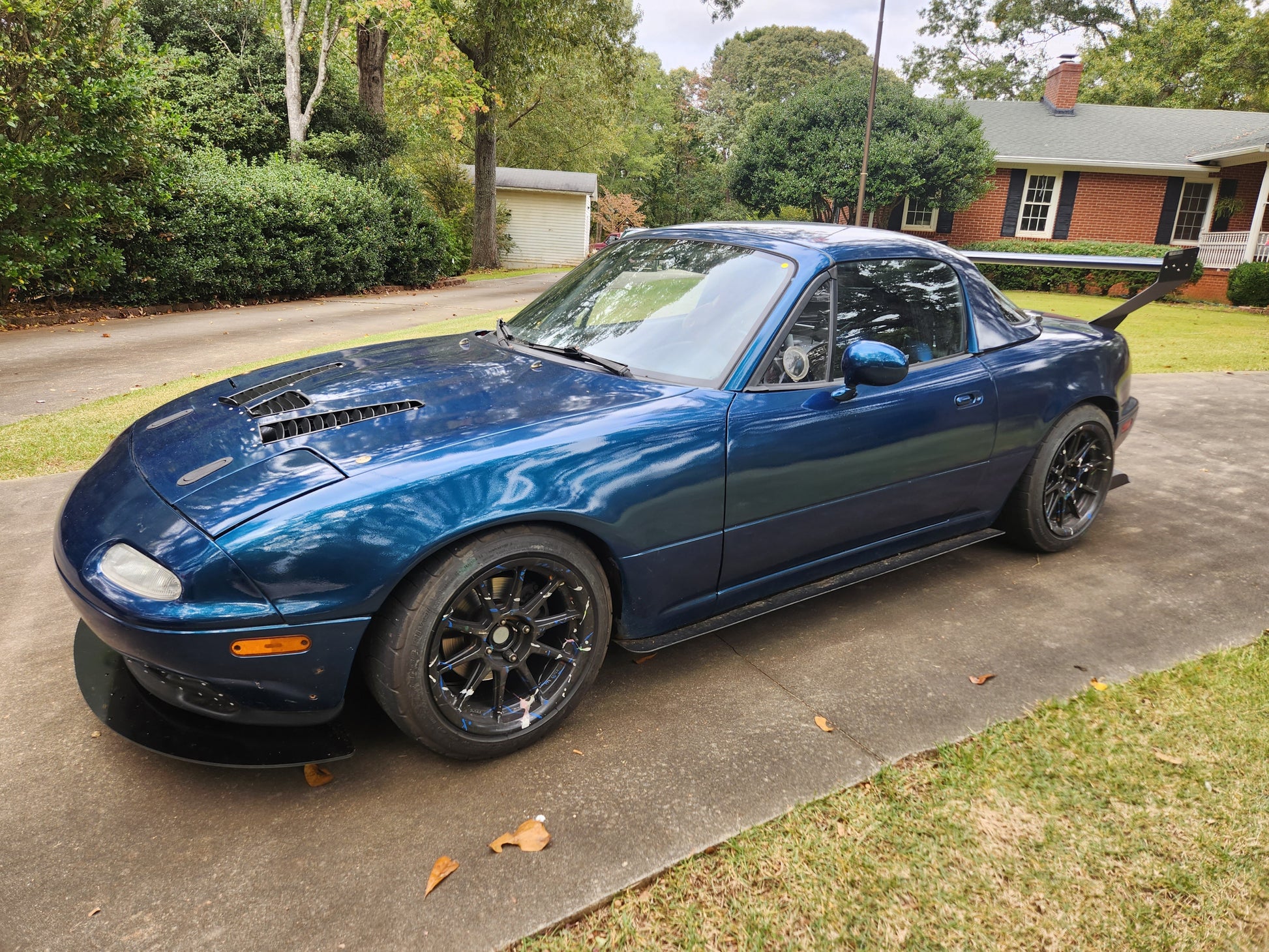 A blue Mazda Miata sports car with black wheels and a custom hood is parked on a concrete driveway in front of a red brick house with a white garage in the background. The car features the 48" Miata V2 Street Wang '90-05 NA/NB by 9livesracing, along with various performance modifications that make it an eye-catching speed demon.