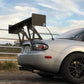 A sleek silver sports car, featuring the Miata Big Wang Kit '06-'15 NC *V1* by 9livesracing, is parked near a white fence. The car boasts black alloy wheels and a soft-top roof. The sky is partly cloudy, with trees visible in the background.