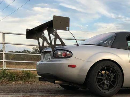 A sleek silver sports car, featuring the Miata Big Wang Kit '06-'15 NC *V1* by 9livesracing, is parked near a white fence. The car boasts black alloy wheels and a soft-top roof. The sky is partly cloudy, with trees visible in the background.