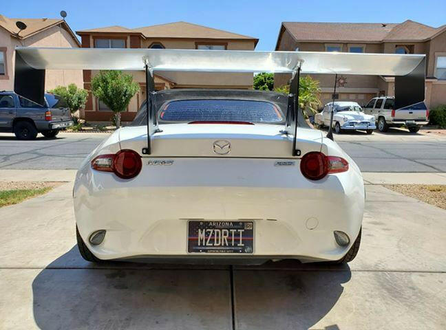 A white Mazda MX-5 Miata is shown from the rear, equipped with the 9livesracing Miata Big Wang Kit 2016+ ND high down-force wing spoiler, parked in a residential area. The license plate reads "MZDRFT1." In the background, there are houses, cars, and a tree on a sunny day.