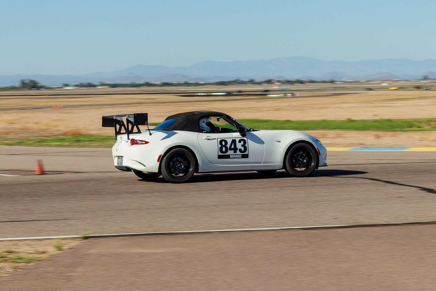 A white Mazda MX-5 Miata sports car, equipped with the 9livesracing Miata Big Wang Kit 2016+ ND and sporting the number 843 on the side, is driving on a racetrack. Featuring a high down-force wing and black wheels, the car cuts through an open area with scattered patches of grass and distant mountains under a clear sky.