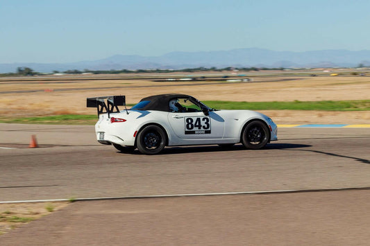 A white Mazda MX-5 Miata sports car, equipped with the 9livesracing Miata Big Wang Kit 2016+ ND and sporting the number 843 on the side, is driving on a racetrack. Featuring a high down-force wing and black wheels, the car cuts through an open area with scattered patches of grass and distant mountains under a clear sky.