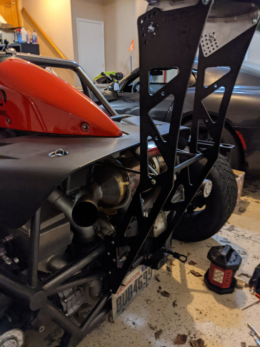 The image depicts a partially assembled Ariel Atom Big Wang vehicle from 9livesracing in a garage. A red motorcycle shell is visible, revealing exposed mechanical components like exhaust pipes and the frame beneath. Nearby, various tools, including a power tool on the floor and a set of steps leading to an upper level, can be seen. The meticulous arrangement indicates attention to lift-to-drag ratio.