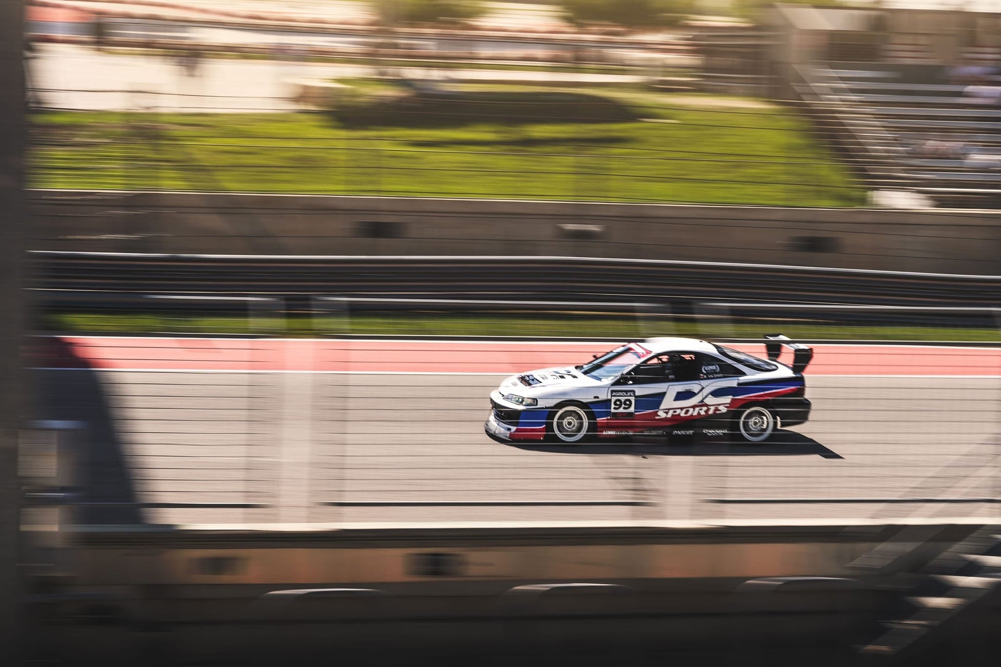 An Integra V2 Big Wang Kit '94-01 DC2 sports car from 9livesracing zooms down the race track, its number 99 emblazoned on the door. The sleek, low-drag design showcases a white body adorned with blue and red stripes, proudly displaying "DC SPORTS" on the side. As the background blurs from high speed, spectators can be seen cheering in the distance.
