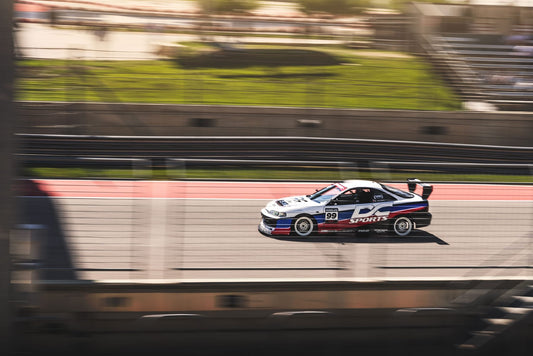 An Integra V2 Big Wang Kit '94-01 DC2 sports car from 9livesracing zooms down the race track, its number 99 emblazoned on the door. The sleek, low-drag design showcases a white body adorned with blue and red stripes, proudly displaying "DC SPORTS" on the side. As the background blurs from high speed, spectators can be seen cheering in the distance.