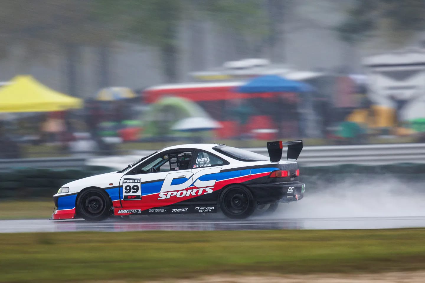 A white and blue sports car with the number 99 on its side races through a wet track, producing splashes of water. The car, equipped for low drag, features a large rear spoiler with endplate mounts from the Integra V2 Big Wang Kit '94-01 DC2 by 9livesracing. The background is blurred with spectators and colorful tents visible in the distance.