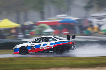 A white and blue sports car with the number 99 on its side races through a wet track, producing splashes of water. The car, equipped for low drag, features a large rear spoiler with endplate mounts from the Integra V2 Big Wang Kit '94-01 DC2 by 9livesracing. The background is blurred with spectators and colorful tents visible in the distance.