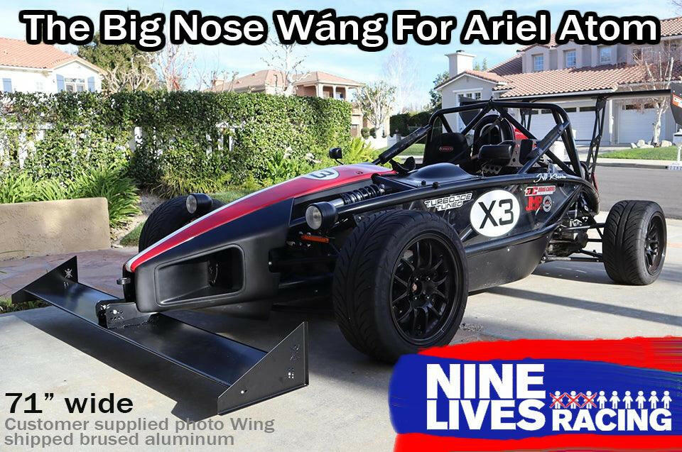 A black Ariel Atom race car with red and white accents sits in a driveway. It boasts a large extruded aluminum front wing, the Ariel Atom Big Nose Wang '00+, measuring 71 inches wide, enhancing its lift-to-drag ratio. Text at the top reads "The Big Nose Wang For Ariel Atom," and the bottom right corner displays the "9livesracing" logo.