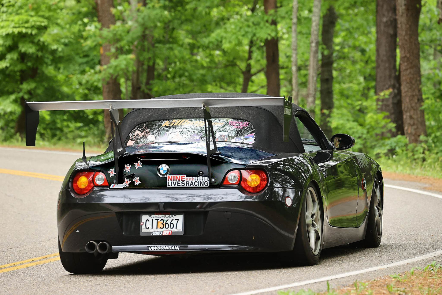 A black BMW Z4 sports car, fitted with the 9LivesRacing Big Wang Kit for 2003-08 models (E85) and adorned with multiple stickers, some of which are placed on the rear windshield, is driving on a winding road surrounded by lush green trees. The license plate reads “CTI 3667,” hinting at its Nine Lives Racing origins.