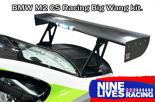 A BMW M2 CS Racing with the Big Wang Kit from 9livesracing is shown featuring a carbon fiber rear wing. The image includes bold text at the top and the 9livesracing logo on the bottom right, emphasizing its exceptional downforce capabilities.