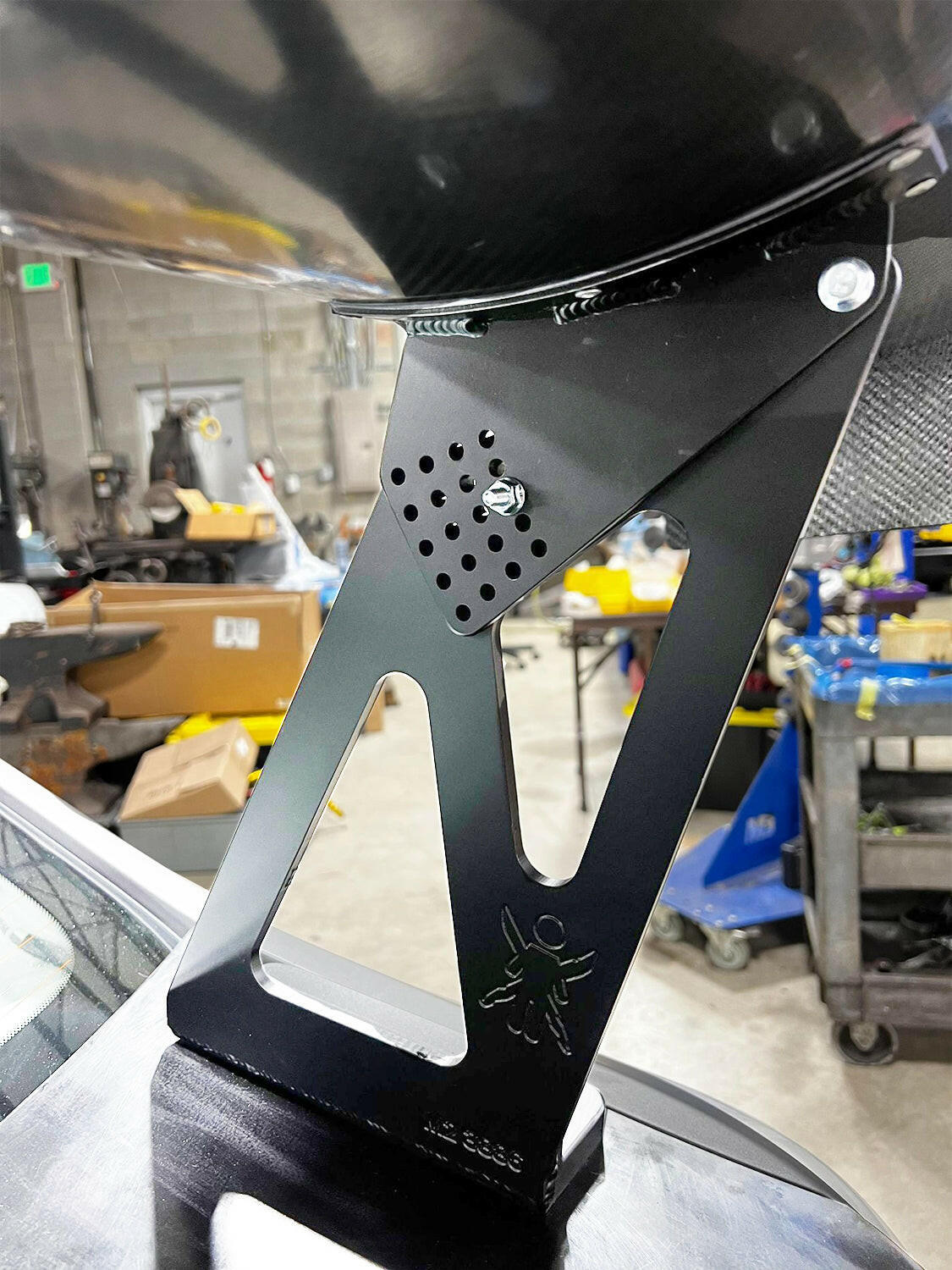 Close-up of a black metal bracket mounted on a surface, with a workshop in the background. The bracket has a perforated area and features the 9livesracing logo with a small ant. Amidst scattered tools and equipment, you notice parts of what seems to be an ambitious BMW M2 CS Racing Big Wang Kit project underway.