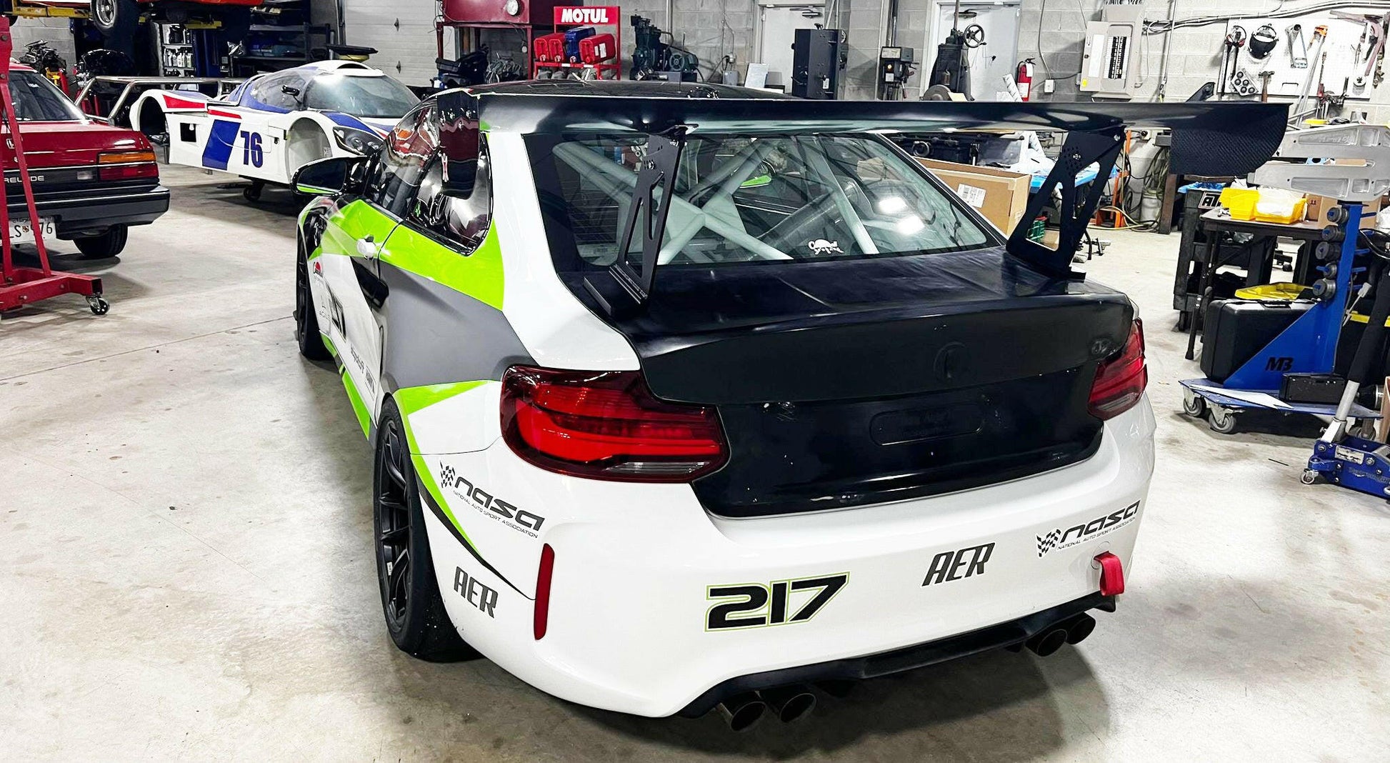 A white BMW M2 CS Racing, accented with green and black details, is parked in a garage. This vehicle features the 9livesracing BMW M2 CS Racing Big Wang Kit for improved down-force and proudly displays the number "217" on its bumper. The presence of other cars and automotive equipment suggests a professional workshop environment.