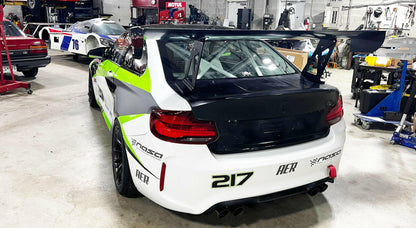 A white BMW M2 CS Racing, accented with green and black details, is parked in a garage. This vehicle features the 9livesracing BMW M2 CS Racing Big Wang Kit for improved down-force and proudly displays the number "217" on its bumper. The presence of other cars and automotive equipment suggests a professional workshop environment.