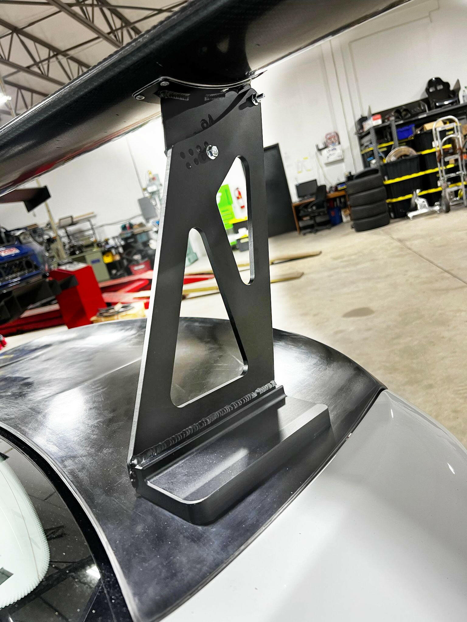 A close-up of an elevated rear spoiler mounted on a BMW M2 CS Racing in a workshop. The robust metal bracket supporting the 9livesracing BMW M2 CS Racing Big Wang Kit is attached to the trunk. The background shows various tools, tires, and equipment, indicating a garage environment.