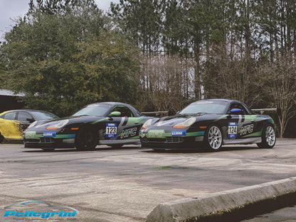 Two Porsche Boxster Big Wang Kit '96-04 986 cars, in green and black with number 123, are parked on a concrete surface in front of tall trees. They feature "WFG" logos and aluminum wings designed for optimal down-force by 9livesracing. A yellow car is partially visible in the background, and the foreground prominently displays a "Pellegrini" logo.