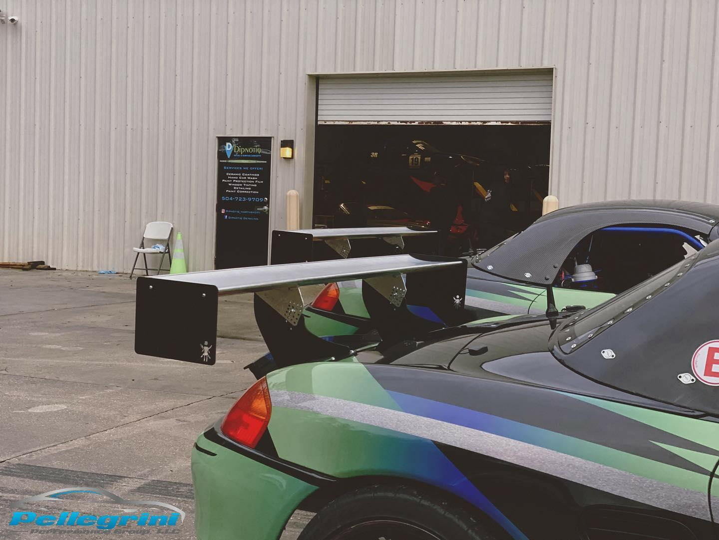 A green and black car featuring the Porsche Boxster Big Wang Kit '96-04 986 by 9livesracing is parked in front of a garage with its door partially open. The background includes a white metal building with a sign and a chair near the entrance. The Pellegrini Performance logo is visible on the ground.