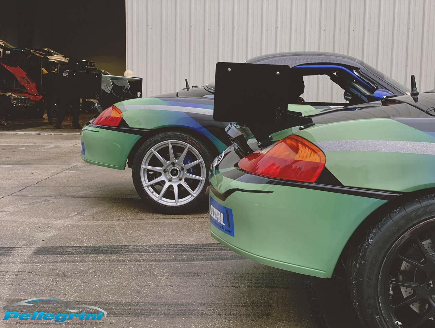 Two Porsche Boxster sports cars from the 1996-2004 series, each equipped with the 9livesracing Big Wang Kit, are parked side by side on a concrete surface near a corrugated metal wall. Both cars, one green and one black, feature sleek designs with aluminum wings and large rear spoilers to maximize down-force. The logo "Pellegrini Performance" is visible on the ground in the foreground.
