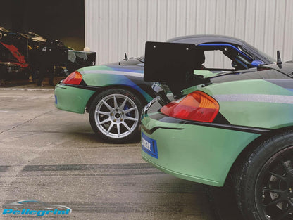 Two Porsche Boxster sports cars from the 1996-2004 series, each equipped with the 9livesracing Big Wang Kit, are parked side by side on a concrete surface near a corrugated metal wall. Both cars, one green and one black, feature sleek designs with aluminum wings and large rear spoilers to maximize down-force. The logo "Pellegrini Performance" is visible on the ground in the foreground.