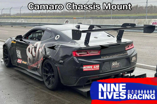 A sleek black Camaro racecar, sporting the number 89 on its side, is parked in a pit area. The car boasts prominent aluminum wings designed to enhance downforce, with mounting points clearly marked by arrows. At the top of the car is text reading "Camaro Wang 2016+ 6th Gen - Chassis Mount," while in the bottom right corner, there's a logo for "9livesracing.