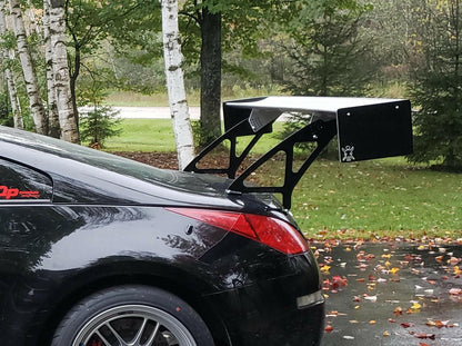 A close-up side view of a black sports car parked on a driveway with a large rear spoiler, which features extruded aluminum wings for optimal 9livesracing 350z Big Wang Kit '02-08 Z33 performance. Behind the vehicle, tall birch trees and scattered autumn leaves create a picturesque autumn backdrop.