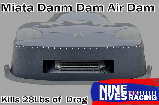 A front view of a grey Mazda Miata with an aftermarket air dam attached. Text at the top reads "Miata Damn Dam '90-05 NA/NB" and at the bottom reads "Kills 28Lbs of Drag". The 9livesracing logo is in the lower right corner, showcasing enhanced downforce.
