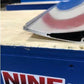 A close-up of a Gurney Flap with a beveled edge, placed on a wooden surface. In the background, there's a blue and white sign with the text "9livesracing" and a small logo of two stick figures.