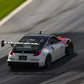 A modified white sports car with a large rear spoiler races on a track, leaving tire marks. The car, equipped with the 9livesracing 350z Big Wang Kit '02-08 Z33 for enhanced down-force and performance, sports racing decals on the side and hood. The blurred background emphasizes the car's motion and speed.