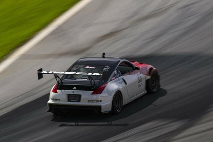 A modified white sports car with a large rear spoiler races on a track, leaving tire marks. The car, equipped with the 9livesracing 350z Big Wang Kit '02-08 Z33 for enhanced down-force and performance, sports racing decals on the side and hood. The blurred background emphasizes the car's motion and speed.
