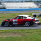 A red and white race car, numbered 96, equipped with a V8 Miata engine and fitted with the Miata Diffuser '90-05 NA/NB from 9livesracing, is speeding on a racetrack. The car features prominent sponsors' logos and a large rear wing alongside its aluminum diffuser. The background shows the racetrack bordered by blue and white barriers and empty grandstands under a clear sky.