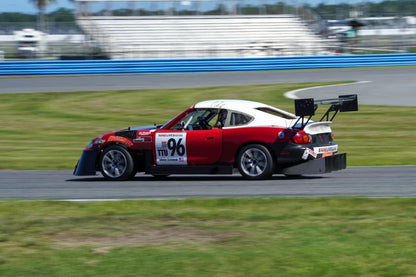 A red and white race car, numbered 96, equipped with a V8 Miata engine and fitted with the Miata Diffuser '90-05 NA/NB from 9livesracing, is speeding on a racetrack. The car features prominent sponsors' logos and a large rear wing alongside its aluminum diffuser. The background shows the racetrack bordered by blue and white barriers and empty grandstands under a clear sky.