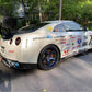 A sleek white sports car adorned with racing decals and the number 14 displayed on the sides and rear is equipped with a GT-R Big Wang Kit '07-22 from 9livesracing, featuring a large extruded aluminum wing for optimal down-force adjustments. It also boasts black wheels with blue rims and numerous sponsor logos on its side. The modified GT3 is parked on a paved driveway, surrounded by lush greenery.