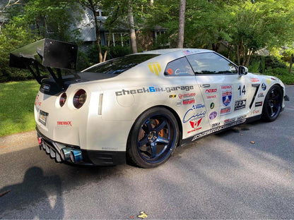A sleek white sports car adorned with racing decals and the number 14 displayed on the sides and rear is equipped with a GT-R Big Wang Kit '07-22 from 9livesracing, featuring a large extruded aluminum wing for optimal down-force adjustments. It also boasts black wheels with blue rims and numerous sponsor logos on its side. The modified GT3 is parked on a paved driveway, surrounded by lush greenery.