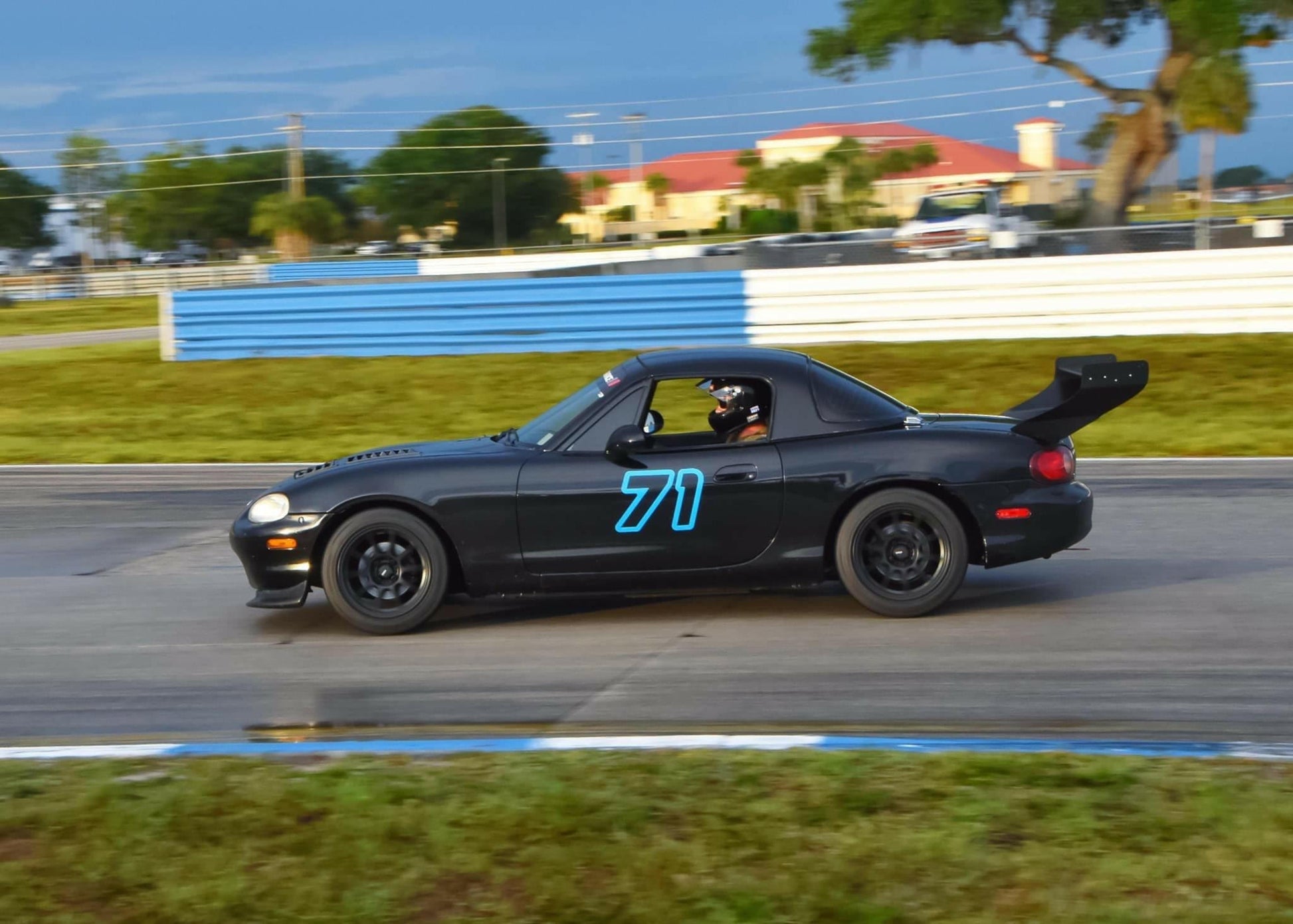 A black Mazda Miata with the number 71 on the door, equipped with a 9livesracing Miata V2 Street Wang '90-05 NA/NB, is racing on a track. The driver, focused and helmeted, maneuvers skillfully. The background reveals green grass, blue and white barriers, and buildings amid trees under a cloudy sky.
