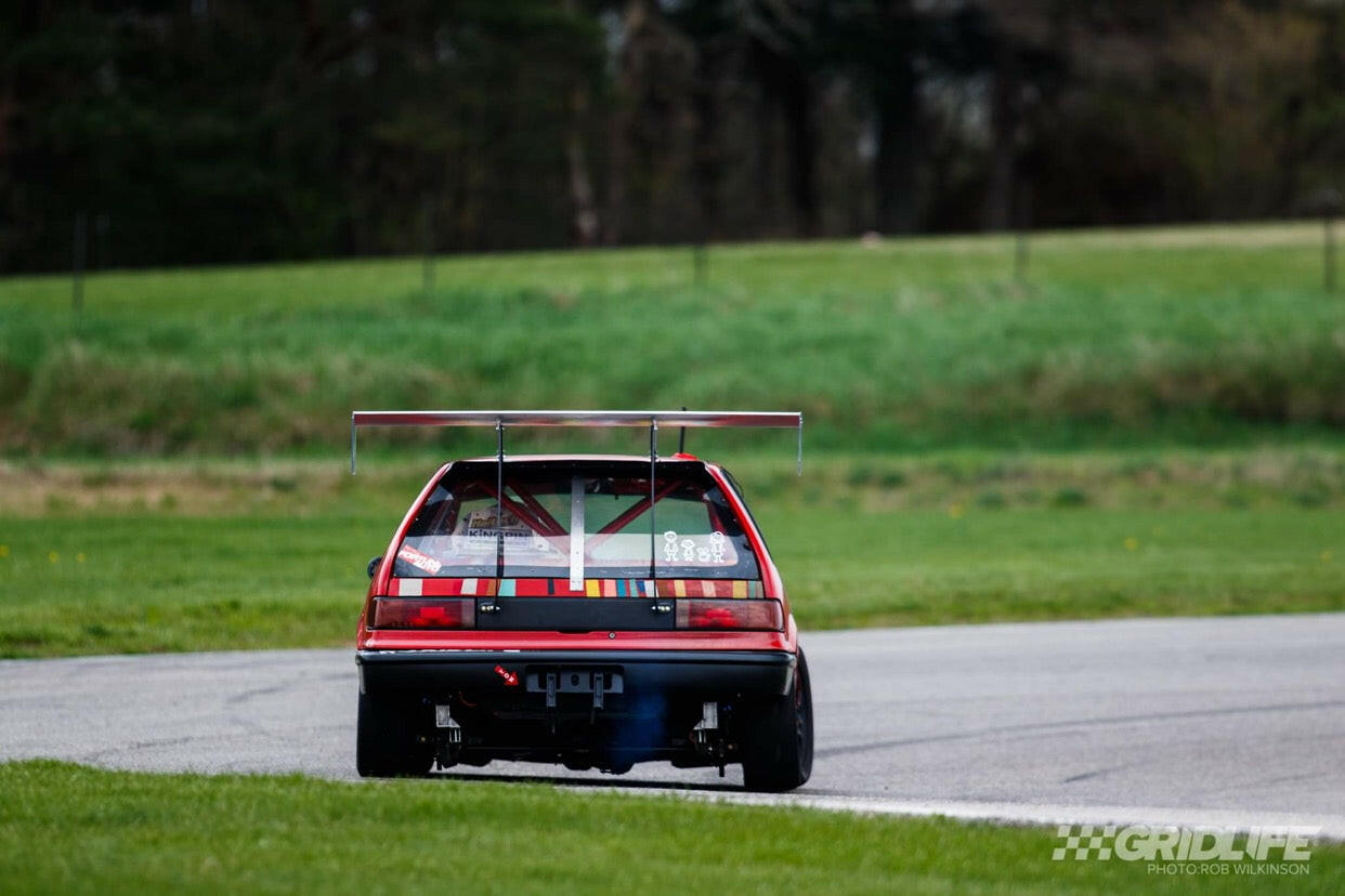 A 9livesracing Civic Hatch EF Wang kit 88-91 in red, equipped with a prominent rear spoiler, cruises on a race track flanked by green grass and trees. The car's rear is adorned with various stickers, its exhaust clearly visible, delivering impressive down-force. The photo is branded with "GRIDLIFE" and the photographer’s name at the bottom right.