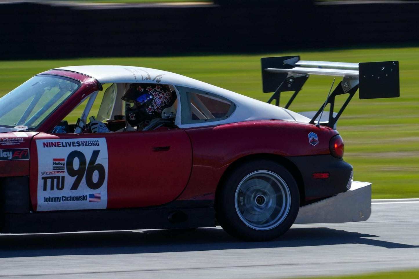A Miata Diffuser '90-05 NA/NB from 9livesracing, installed on a red and white race car with the number 96 on the door and an aluminum rear wing, is speeding on a track. The V8 Miata displays visible sponsor decals. The driver, partially visible through the window and wearing a helmet, adds to the dynamic scene as the blurred background emphasizes their motion.