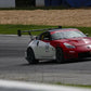 A red and white race car with the number 50 on the door speeds around a racetrack, equipped with a 9livesracing 350z Big Wang Kit '02-08 Z33, featuring GT3 performance and extruded aluminum wings. The car boasts a prominent rear spoiler and various sponsor logos. The track is lined with grass and barriers, with tire walls and gravel in the background.